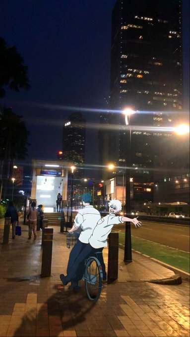 An anime-style illustration of two students in white shirts riding a bicycle together down a city street at night, with one reaching their arm out joyfully toward the illuminated skyscrapers.