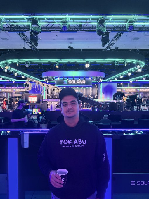 A man wearing a black 'TOKABU - THE SPIRIT OF GAMBLING' hoodie and cap holding a cup, standing in front of a stage lit with purple and blue lights displaying 'SOLANA' branding.