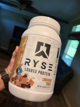 A hand holds up a tub of RYSE Loaded Protein in the flavor "Cinnamon Toast," featuring a 9G graphic on the label. This fits the "GM! #HealthGoals" post by showcasing a popular protein supplement as part of a fitness-focused lifestyle.