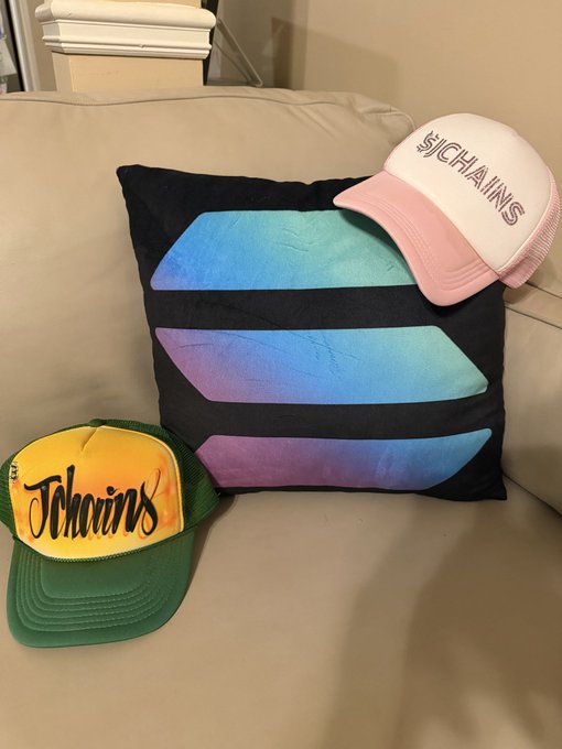 Two trucker hats branded with the "Jchains" logo—one in green and yellow, the other in white and pink—rest on a couch alongside a black pillow featuring colorful iridescent geometric shapes, with the hats displaying the $JCHAINS ticker referenced in the post.