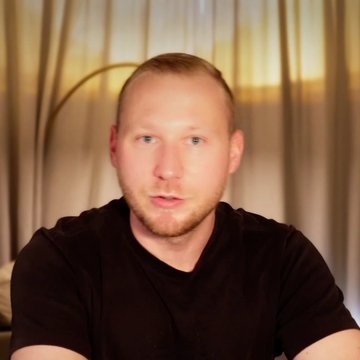 A selfie-style photo of a man with short blonde hair wearing a black t-shirt, sitting indoors with curtains and warm lighting in the background.