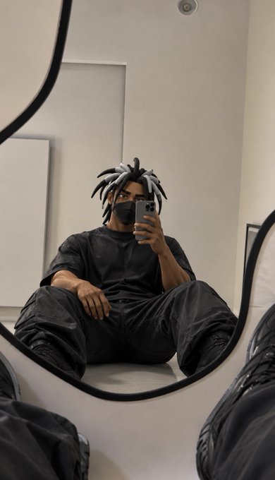 A mirror selfie of a person with black and white dreadlocks wearing a black face mask and all-black outfit, sitting down while taking the photo with their phone.