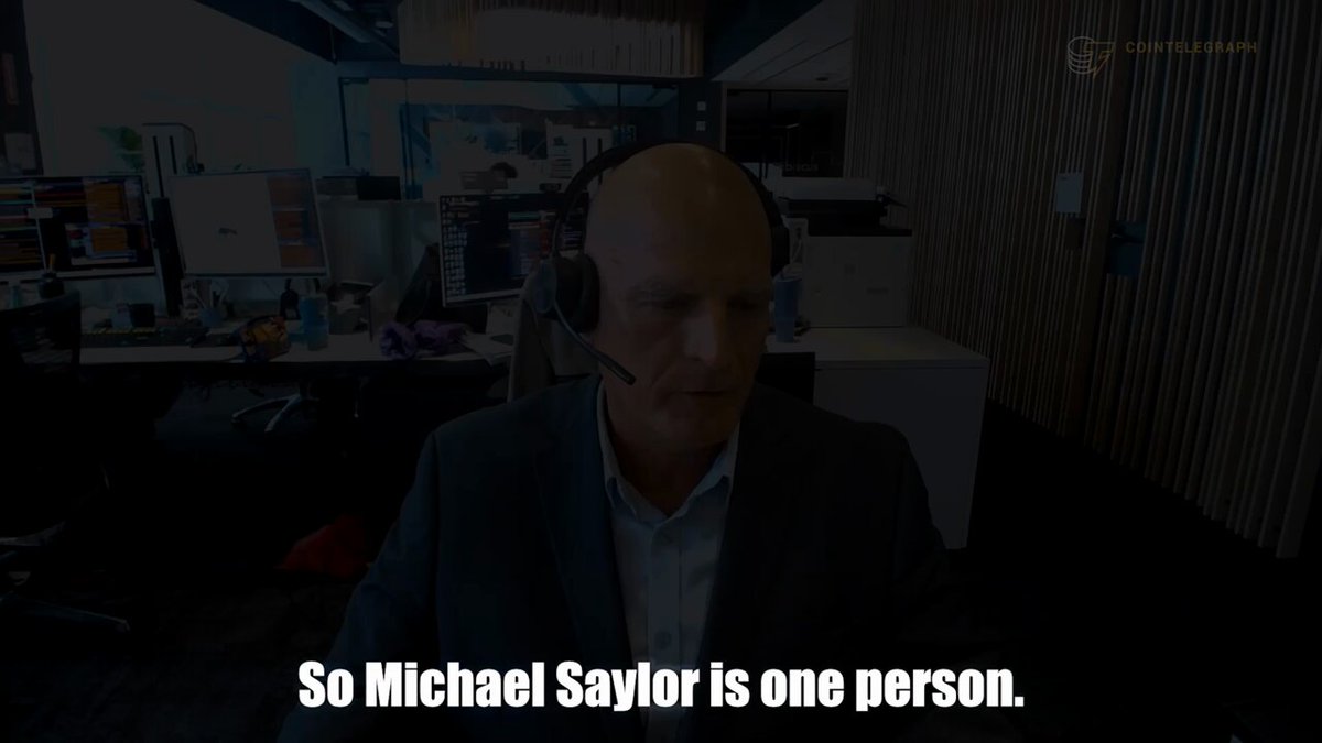 The image shows an office setting with multiple people present, including one man wearing headphones who appears to be a businessman or a tech professional. The background features several monitors and computers, indicating that this could be a workspace for a company or an individual working in technology or finance. The main focus of the image is on the person wearing headphones sitting at their desk, with text overlaying the scene that reads "So Michael Sawyer is one person." This suggests that the image might be related to a discussion about the man's job title or his role within the company.