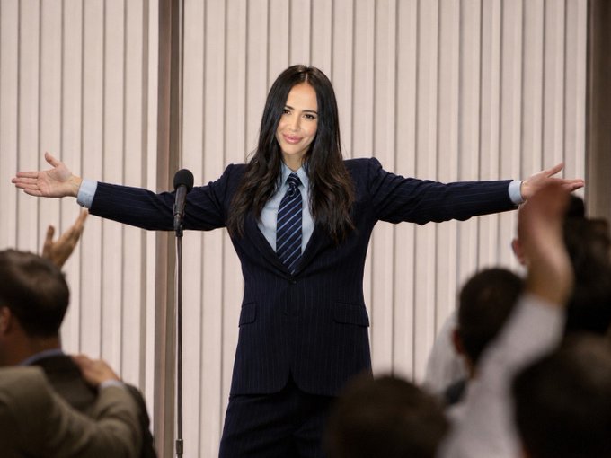 A woman in a pinstripe suit and tie stands with arms outstretched at a microphone, appearing to address a crowd in a triumphant pose. The image conveys confidence and rallying energy, fitting the post's declaration of unwavering commitment to crypto despite volatility.