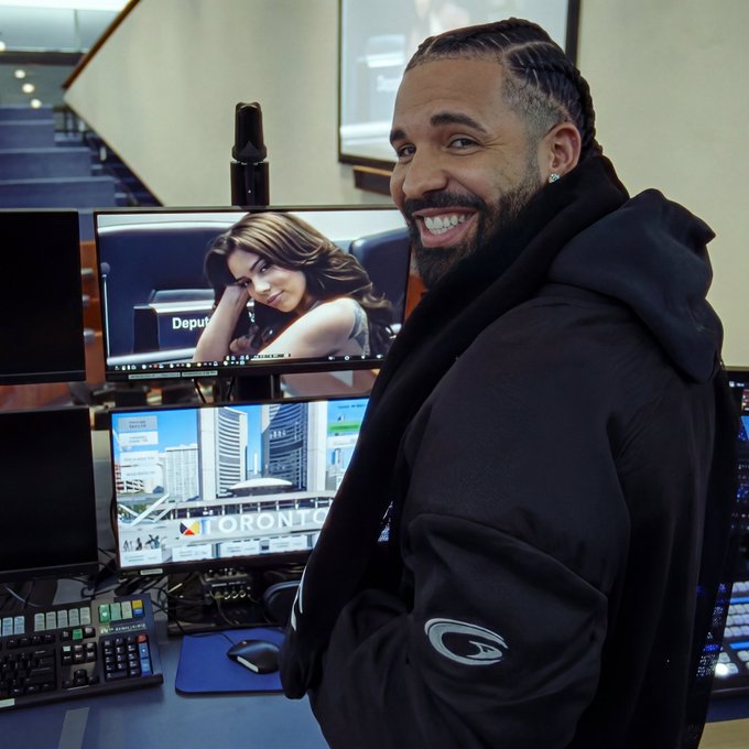 A smiling man in a black Gatorade jacket and black scarf stands in front of computer monitors displaying a photo of a woman and a Toronto cityscape.
