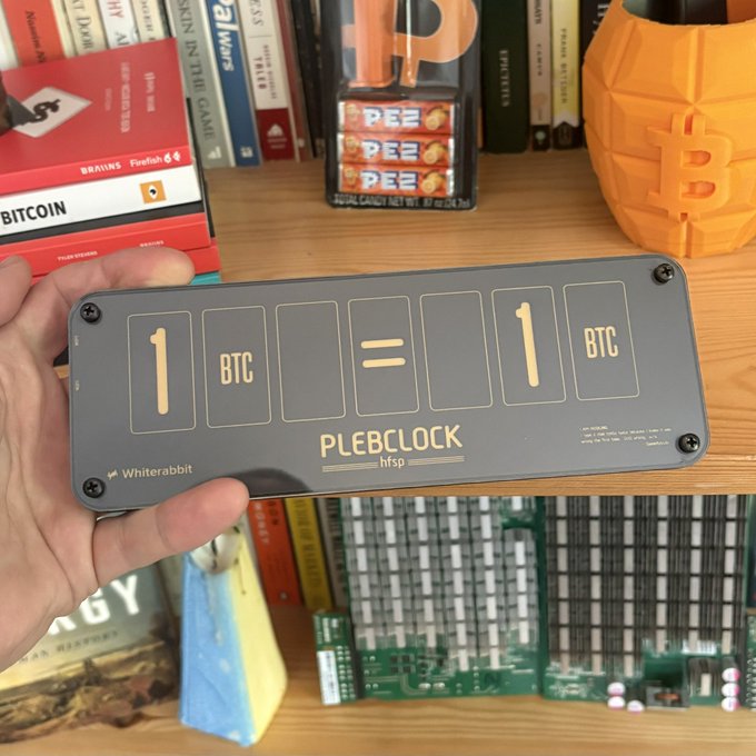 A hand holding a dark grey 'PLEBCLOCK hfsp by Whiterabbit' physical Bitcoin split-flap display showing '1 BTC = 1 BTC', with PEZ dispensers, books including 'MASTERING BITCOIN', and a Bitcoin-themed object visible on the desk in the background.