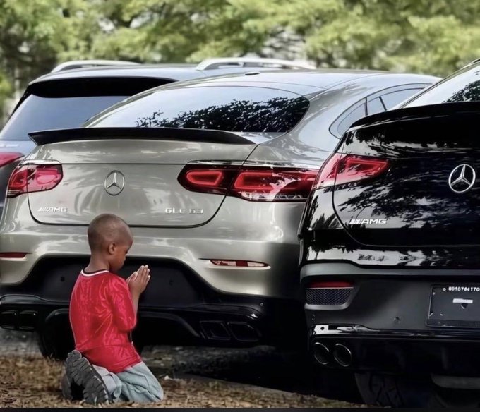 A young boy kneels in prayer in front of two parked Mercedes-AMG SUVs, one gray and one black.