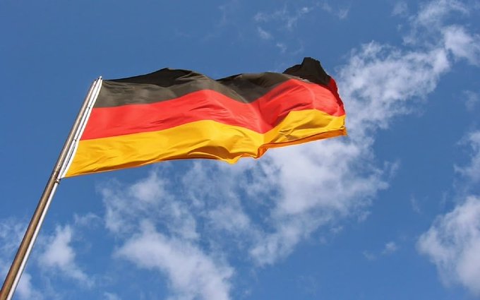 The German national flag with its black, red, and gold horizontal stripes waving against a blue sky with scattered white clouds.