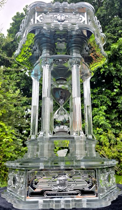 An ornate, decorative glass hourglass sculpture with columns and intricate details, photographed outdoors with green foliage in the background.