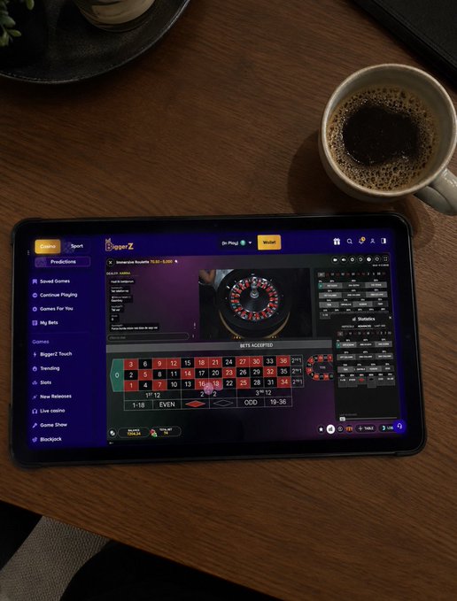 A tablet displays the BiggerZ live casino platform showing Immersive Roulette gameplay, with the betting layout clearly showing the standard 1-36 numbers plus 0 and 00, while a coffee mug sits on the wooden table nearby—perfectly matching the post's relaxed setup of enjoying coffee and playing their favorite game using the promo code ZENIA via the referral link.