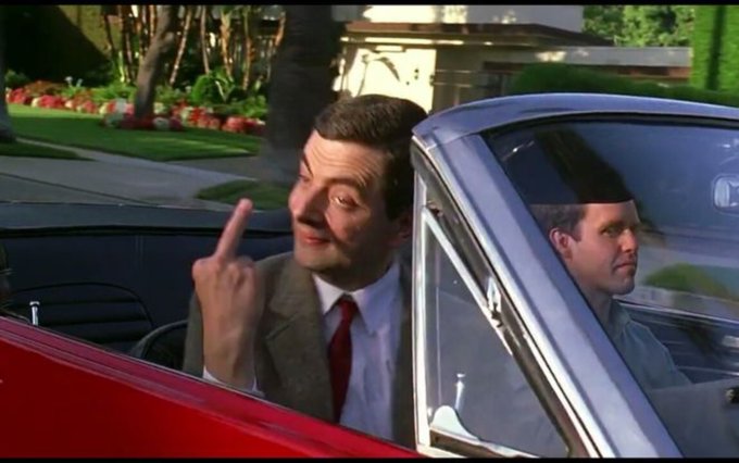 A man in a suit sitting in the passenger seat of a red car makes an obscene gesture with his middle finger.