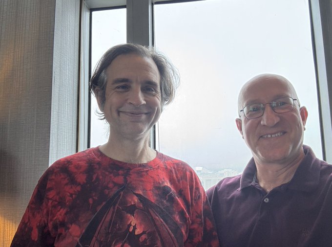 Two men pose together in front of a large window with a city view, including the man referenced as @zooko on the left wearing a red patterned shirt. The photo captures a warm, friendly meeting likely between Zooko Wilcox (creator of Zcash) and a friend or colleague.
