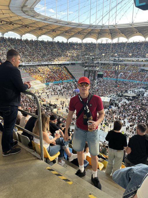 The image shows a man wearing a red shirt standing at the top of a staircase, holding a drink. He appears to be enjoying his time at an outdoor event or stadium. The scene also includes several other people scattered around the area, some sitting on chairs while others stand and socialize. A cup can be seen in the foreground as well.