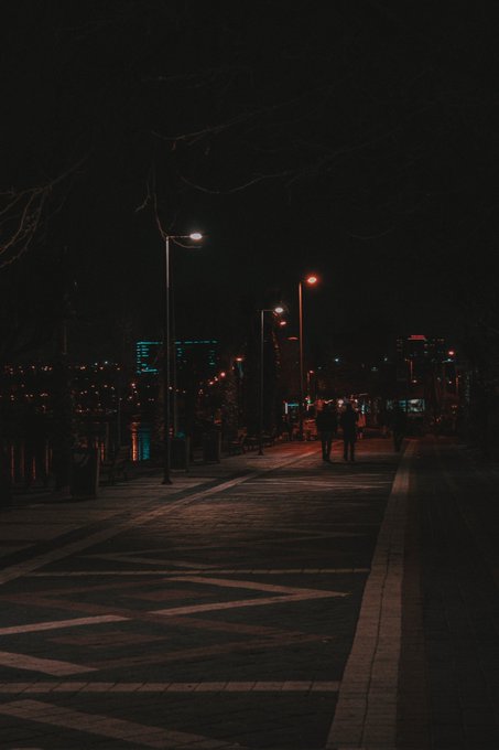 The image shows a couple walking down a sidewalk at night, with street lights illuminating their path. There are also several other people visible in the scene, some of whom appear to be carrying backpacks or handbags. The overall atmosphere suggests that this is an urban setting where people are out and about during nighttime hours.