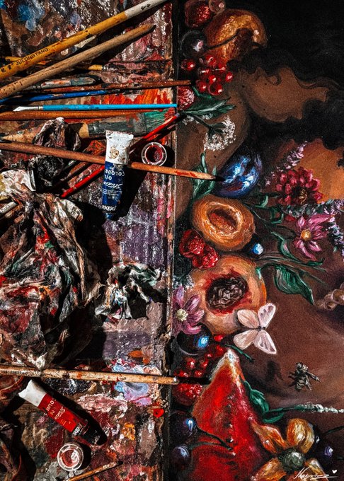 An overhead view of an artist's workspace featuring scattered paintbrushes, paint tubes, and crumpled rags on the left, alongside an in-progress still life painting of fruits, flowers, and insects on the right.