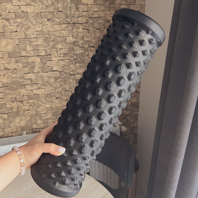 A person wearing a beaded bracelet holds up a black textured foam roller with a bumpy, spiky surface against a brick wall background.
