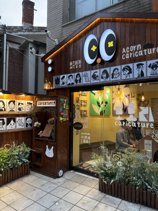 The storefront of 'Acorn caricature' featuring a wooden facade, a logo with large eyes, and a sign decorated with various character drawings of patrons.
