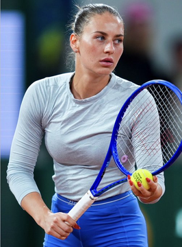 A female tennis player in a light gray long-sleeve top and blue skirt holding a tennis racket and a yellow ball, appearing focused mid-game.