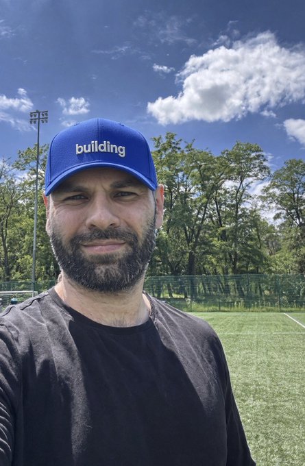 A man wearing a blue cap with 'building' text and a black t-shirt takes a selfie outdoors on a green sports field with trees and a stadium light in the background.