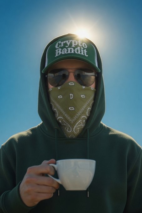 A person wearing a green hoodie, a green bandana over their mouth, dark sunglasses, and a green trucker hat reading 'Crypto Bandit' holds a white teacup against a bright blue sky.