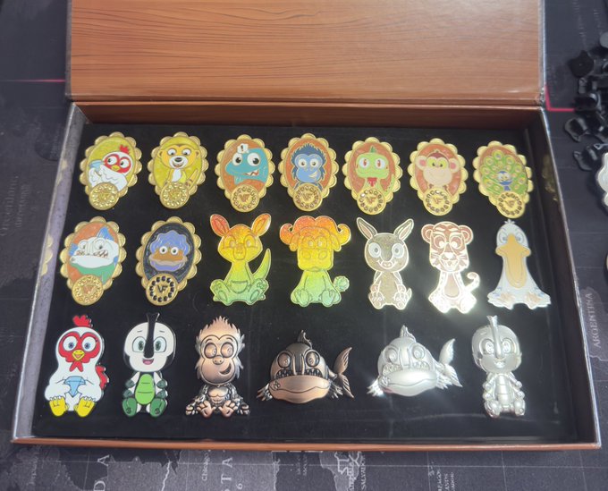 The image shows a collection of 16 different pins, each featuring unique designs and colors. These pins are neatly arranged inside an open wooden box or display case, showcasing their various shapes and sizes. The text "A few more @veefriends pins added" suggests that there might be additional pins in the collection, possibly to add variety or enhance the visual appeal of the display.

The image also contains a small portion of a black background with white text, which could potentially be part of an online store description or product information.