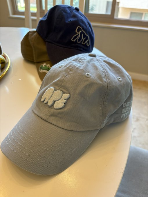iced gray baseball cap with white "ape" logo, accompanied by a brown and blue hat on the table.
