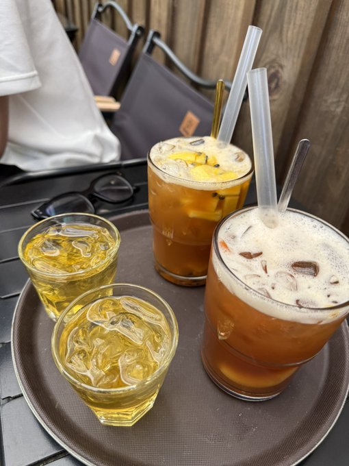 A tray holds two iced amber-colored drinks with glass straws and two small glasses of yellow liquid, set on an outdoor table beside a pair of glasses.