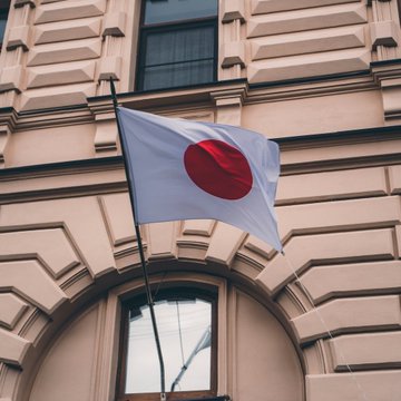 The image shows the flag of Japan — a white banner with a central red circle — displayed outside a building with classical architectural details, symbolizing the country whose Financial Services Agency (FSA) is tightening crypto exchange withdrawal rules to combat scam-related fund transfers.