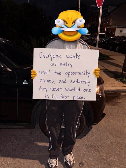 A person wearing an angry emoji filter holds a handwritten sign reading 'Everyone wants an entry until the opportunity comes, and suddenly they never wanted one in the first place.'