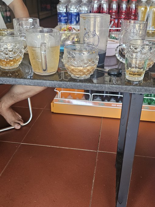 A glass table holds various drinking glasses, pitchers, and bottles of beverages, with a person's bare foot visible underneath on a tiled floor.