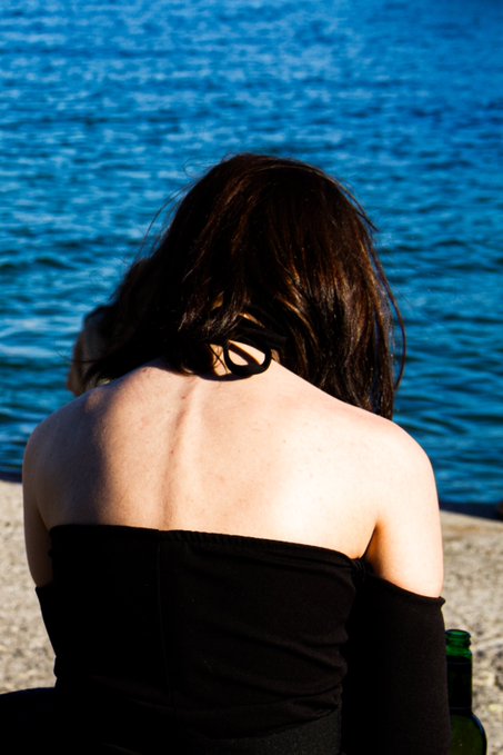 A photograph of a woman with dark hair, wearing a black off-shoulder top, sitting with her back to the camera and looking out at blue water.
