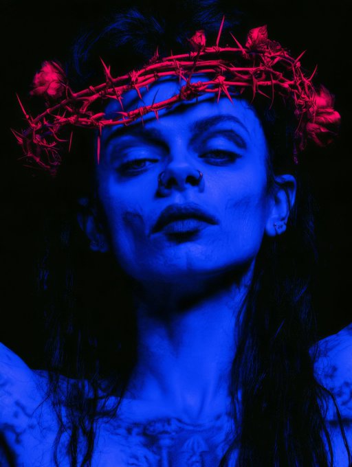 A woman with closed eyes wears a crown of red glowing thorns against a dark background, illuminated by striking blue light.
