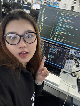 A young woman with glasses points toward the screen while sitting in front of two monitors displaying programming code.