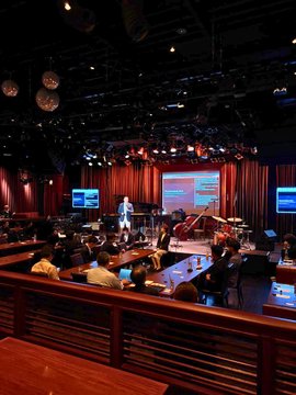 Photograph of a conference or panel discussion taking place in a dimly lit venue with wooden architectural elements, where speakers are presenting on stage to an audience seated at tables with monitors.