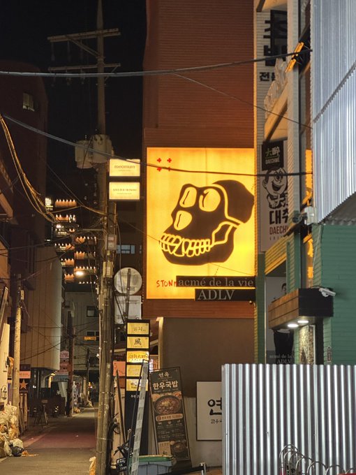 A nighttime street scene featuring a bright yellow illuminated billboard with a black skull design and the text 'STONHAMC DÉ LA VIE ADLV', alongside other Korean signage on a narrow urban alleyway.