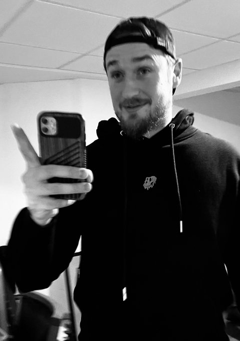 A black and white selfie of a bearded man wearing a backwards cap and a hoodie with a logo. He's smiling while holding up a phone with a textured case.