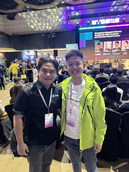 Two men wearing event lanyards pose for a photo at what appears to be the MYBASE event, with a stage presentation titled 'Navigating Malaysia's Startup Ecosystem: What Investors and Founders Need to Know' visible in the background.