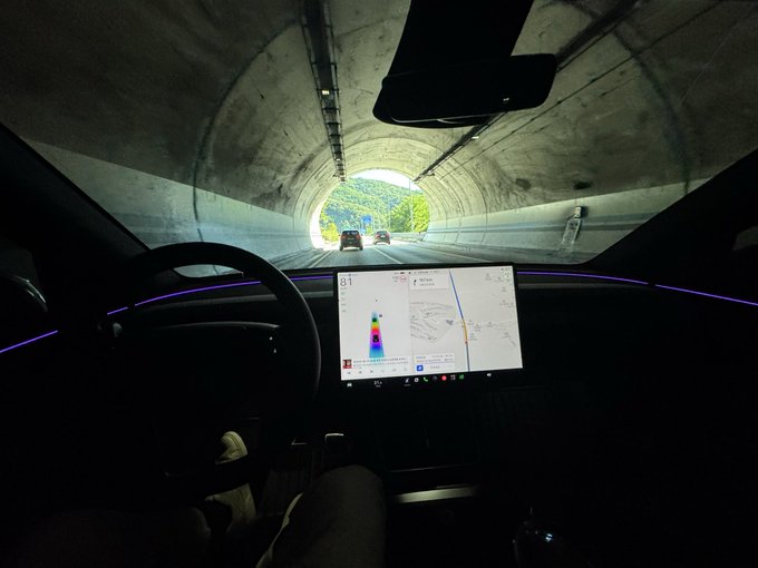 A view from inside a Tesla driving through a tunnel, showing the dashboard screen with a map and navigation interface illuminated against the dark interior, with purple ambient lighting strips visible on the door panels.