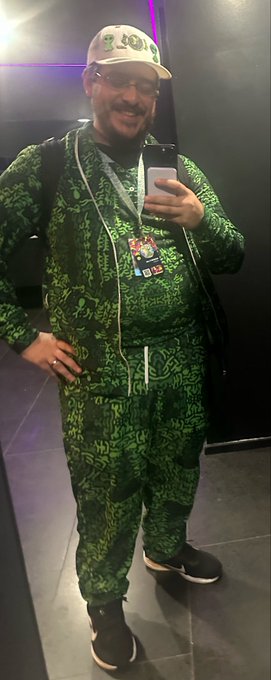 A man takes a mirror selfie while wearing a matching green and black patterned hoodie and pants, a white cap with green designs, glasses, and black sneakers, posing with one hand on his hip while holding his phone. The image captures his enthusiastic outfit at the VeeFriends TCG event, as referenced in the post where he mentions enjoying the championship despite not winning.