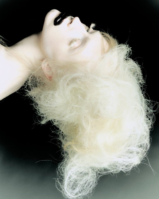 A high-contrast, artistic portrait of a person with dark lipstick and voluminous, backlit blonde hair, posed with their head tilted backward against a dark background.