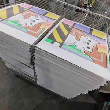 A stack of printed collectible cards featuring a cartoon character with bunny ears is displayed in a print shop, awaiting production. The image relates to the post by showcasing the physical printing process of NFT-inspired merchandise, likely tied to the @elmonx_official project, bridging digital collectibles with tangible products.