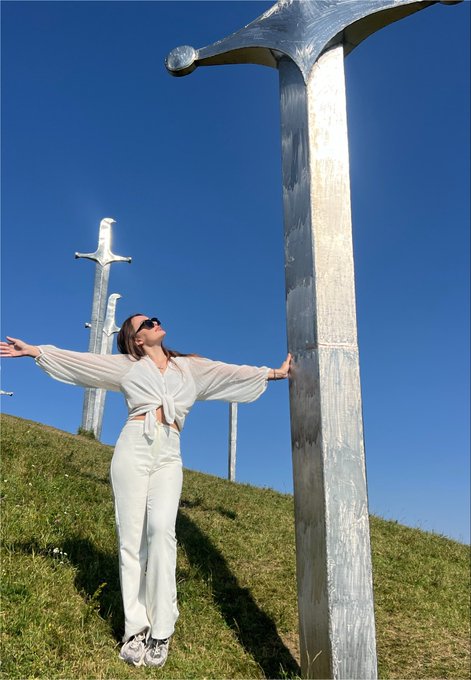 A woman wearing a white tie-front top and white pants stands with arms outstretched on a grassy hill among large silver cross sculptures against a clear blue sky.