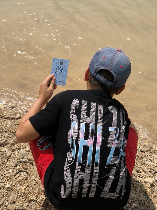 A person wearing a blue cap and a black t-shirt with "CHILL SHIT" printed on the back sits on a rocky riverbank, holding up a small blue card.