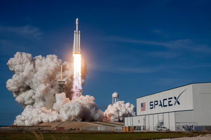 A SpaceX rocket launches from a pad next to a building prominently displaying the "SPACEX" name and American flag, symbolizing the company's high-profile rise as its shares become a major driver of tokenized equity trading activity following its public market debut.