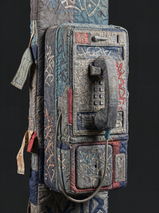 A sculptural artwork resembling an old payphone mounted on a fabric-covered post, crafted from quilted and stitched textiles in muted blue and gray tones with graffiti-style markings.