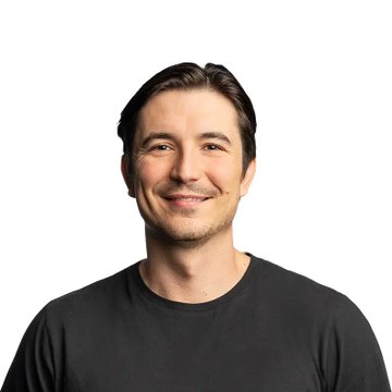 A portrait of a smiling man with short dark hair and stubble, wearing a plain black t-shirt against a white background.