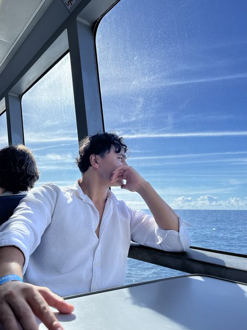 A man in a white linen shirt sits by the window of a ferry or boat, gazing thoughtfully out at the ocean and blue sky.