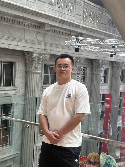 A man poses in a white t-shirt inside a grand hall featuring classical stone columns and glass railings, with a red banner visible in the background. The casual, confident photo aligns with the post's playful "thin skin theory" challenge directed at CZ Binance, suggesting the man is gearing up for the experiment.