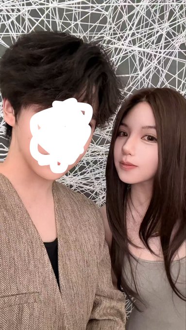 A selfie of a young couple standing close together; the man on the left has his face obscured by a white flower sticker, while the woman on the right has long dark hair.