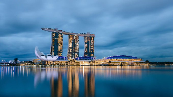 A twilight photograph captures the iconic Marina Bay Sands hotel in Singapore, with its three towers and rooftop SkyPark reflected in the water below.